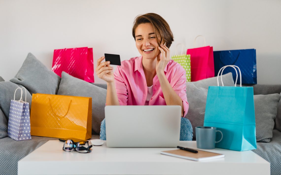 happy smiling woman in pink shirt on sofa at home among colorful shopping bags holding credit card paying online on laptop on sale discount from home social distancing self isolation